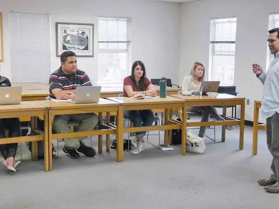 Tory Fodder stands to the right of the image in a blue and white checkered button-up shirt. Toward the left are four students sitting in a single row at yellow-hued wooden desks. Three of the four have silver laptops open in front of them. 