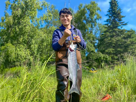 Torin Jacobs II poses in waders with a large fish, likely a Chinook salmon, on a sunny day in Alaska.