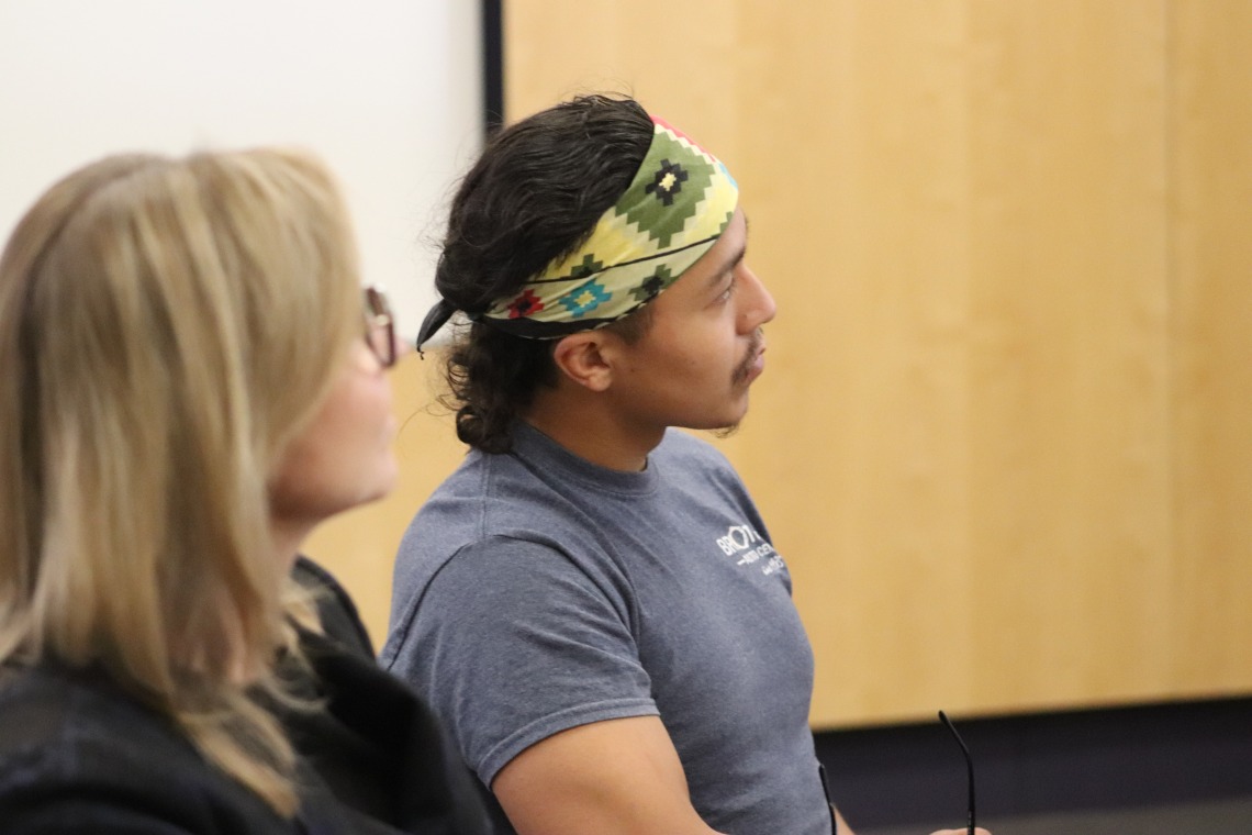 Adam Fernandez, wearing a yellow bandana as a headband, watches an Indigenous Data Sovereignty talk at the BIO5 Insititute with Jennifer Barton in the foreground,