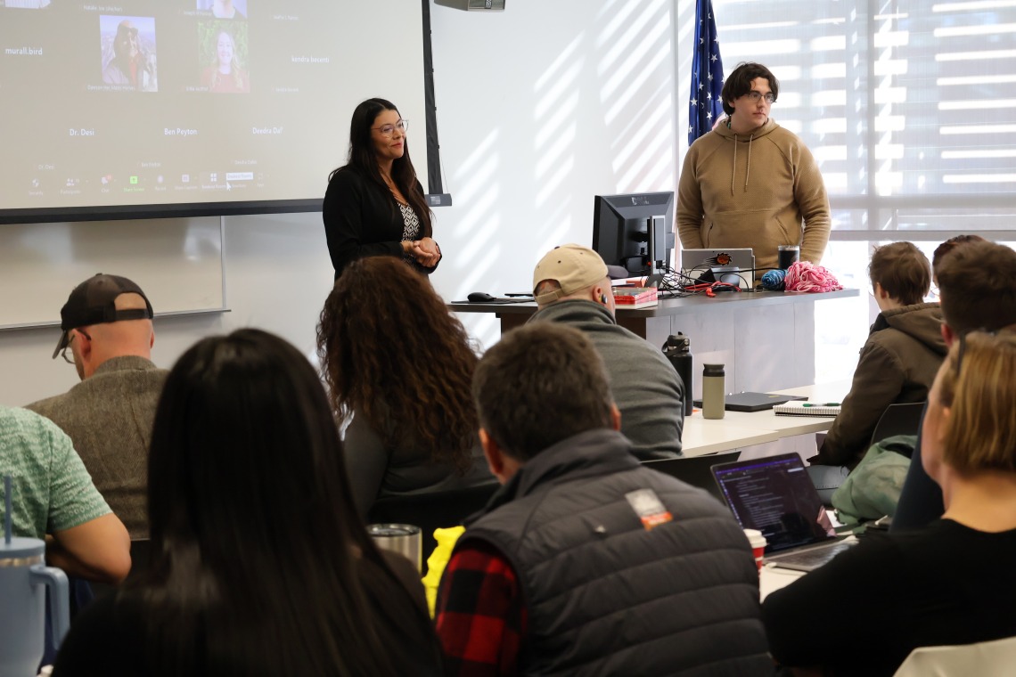 Desi Smal-Rodriguez smiles in front of a full classroom with a Zoom grid of virtual attendees projected behind her. To her left, PhD student William Carson stands behind a desk ready to help.