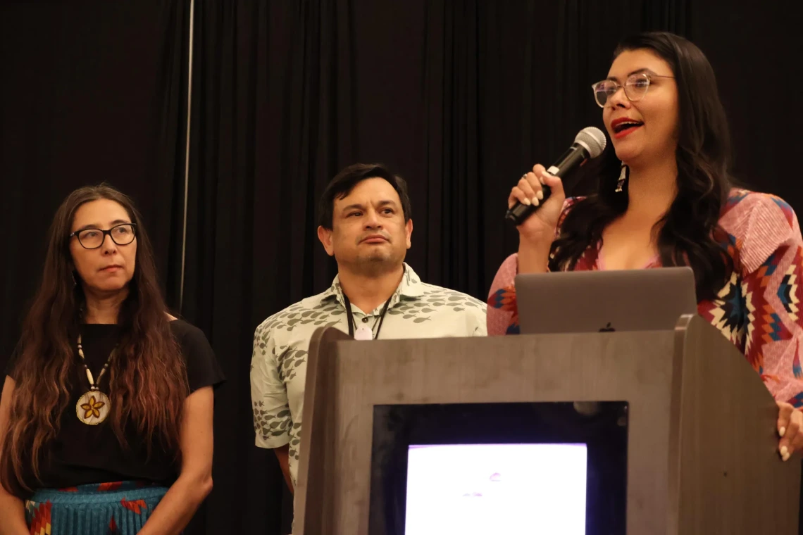 Desi Small-Rodriguez speaks behind a podium as Stephanie Russo Carroll and Randy Akee look on from behind