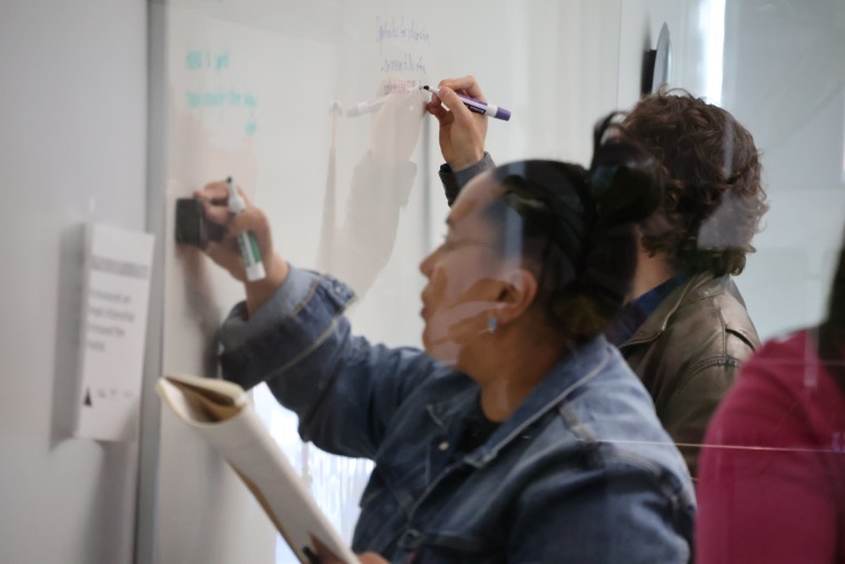 Two students write on a whiteboard during a JIT class. A glare can be seen on the glass between the camera and the subjects.