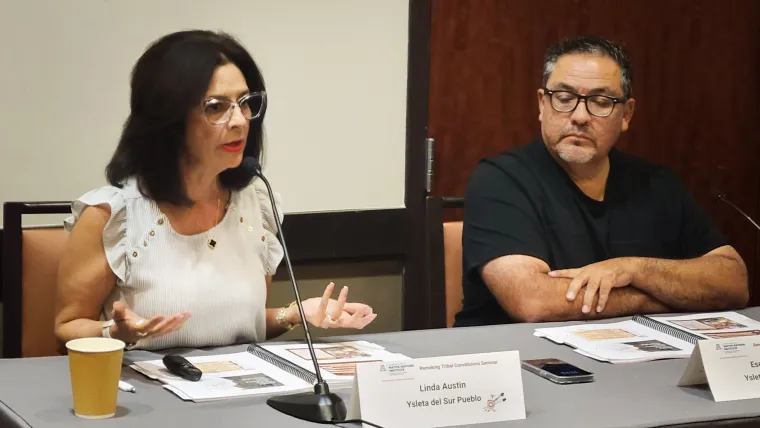 Linda Austin and Zeke Garcia sit behind a gray table with black table microphones and folded namtages in front of them.