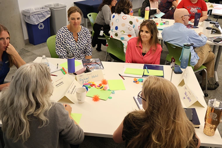 Seminar attendees discuss as a group while sitting around a rectangular table covered in neon post it notes, markers, and other materials