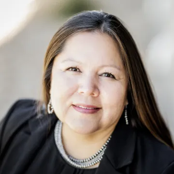 A portrait of Candi Running Bear in a black jacket and shirt. She wears a multi-strand silver necklace and sliver hoop earrings.