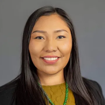 A portrait of Melissa Wheeler against a gray background. She wears a turquoise necklace, black jacket and dark green textured shirt.