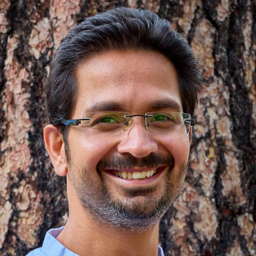 A portrait of Pranay Ranjan in front of the broad trunk of a pine tree. He wears frameless square glasses and a short beard,
