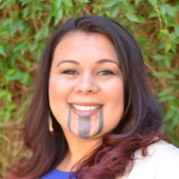 A portrait of Seafha Ramos in a blue shirt and white cardigan. Dr. Ramos wears gold earrings and tribal tattoos on her chin.