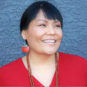 Elisha smiles in a red v-neck top, dangling red earrings and a pair of beaded necklaces in front of a blue stucco wall.