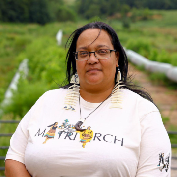 Crystal Cavalier-Keck wears glasses, large earrings shaped like fish skeletons, a white t-shirt that reads "Matriarch" and a necklace with a curved brown pendant as she stands in front of what looks like an agricultural field.