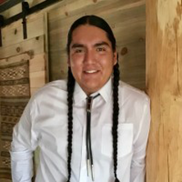 Damon Clark wears a white button-up shirt, braids and a bolo tie as he leans against a sanded wooden pillar in this indoor photo taken inside what looks like a log cabin.