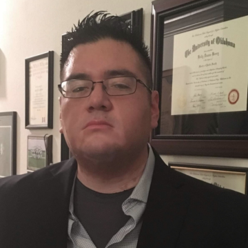 Kelly Berry wears thin-framed glasses, spiked hair, a black jacket, blue collared shirt and dark gray undershirt as he frowns at the camera in front of a wall of degrees and other framed documents.