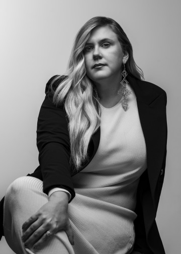 Black and white photo of producer Kaitlin Mooney in a light-colored dress and dark blazer against a gray background.