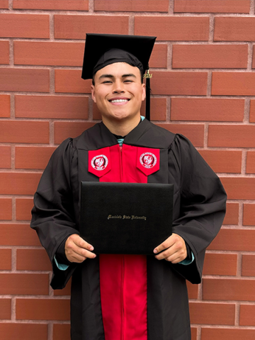 Jacobs wears a graduate cap and gown and holds his degree from Montclair State while standing in front of a red brick wall.