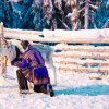 A man in traditional Indigenous Saami clothing kneels in the snow next to a reindeer.